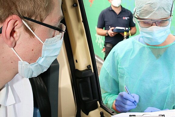 Ein junger Mann mit einer blauen Maske schaut zu einer Gesundheitshelferin, die in einer Schutzkleidung und einer blauen Maske steht. Sie hält einen Stift und notiert Informationen. Im Hintergrund ist ein weiterer Helfer sichtbar, der ebenfalls an einem medizinischen Einsatz beteiligt ist.