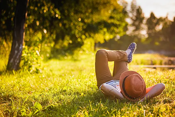 Eine Person liegt entspannt auf einer sonnigen Wiese im Park, Beine übereinandergeschlagen, Strohhut über dem Gesicht. Warmes Abendlicht, Bäume im Hintergrund. Szene vermittelt Ruhe und Sommeridylle.