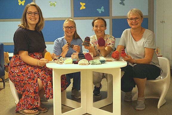Vier Frauen sitzen um einen Tisch und halten handgefertigte, gestrickte Mützen in ihren Händen. Auf dem Tisch liegen weitere Mützen in verschiedenen Farben. Die Atmosphäre wirkt freundlich und kreativ, mit einer hellen Umgebung und bunten Dekorationen im Hintergrund.