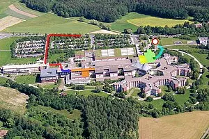 Luftbild einer Anlagenübersicht mit Gebäuden, Parkplätzen und umliegenden Grünflächen. Rote und grüne Pfeile weisen auf verschiedene Wege hin, während ein roter Punkt einen spezifischen Standort markiert. Die Umgebung ist ländlich mit Wäldern und Feldern im Hintergrund.