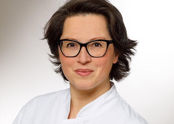 Portrait einer Frau mit kurzen, dunklen Haaren und Brille. Sie trägt eine weiße Kitteljacke und lächelt freundlich. Der Hintergrund ist hell und neutral, was den Fokus auf ihr Gesicht lenkt.