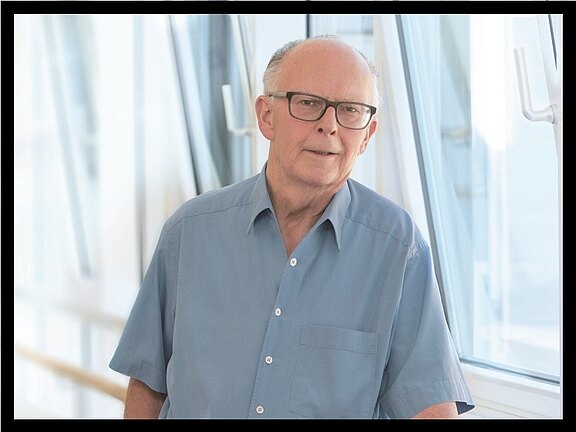 Ein älterer Mann mit Brille steht lächelnd in einem hellen Innenraum. Er trägt ein blaus Hemd und lehn sich leicht an eine Wand neben einem Fenster. Der Hintergrund ist unscharf und zeigt eine moderne Architektur mit viel Licht.