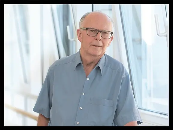 Ein älterer Mann mit Brille steht lächelnd in einem hellen Innenraum. Er trägt ein blaus Hemd und lehn sich leicht an eine Wand neben einem Fenster. Der Hintergrund ist unscharf und zeigt eine moderne Architektur mit viel Licht.