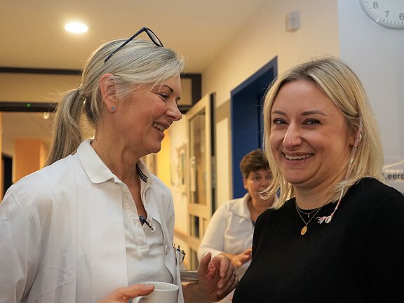 Zwei Frauen lachen und sprechen in einer Klinik. Links eine Frau im weißen Kittel mit Brille auf dem Kopf und einer Tasse in der Hand, rechts eine jüngere Frau im schwarzen Oberteil. Die Szene wirkt freundlich und entspannt.