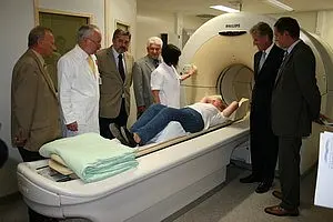 Eine Gruppe von Fachleuten steht um einen Patienten, der auf einer Liege liegt und sich einem MRT (Magnetresonanztomographie) unterzieht. Der Raum wirkt medizinisch und gut ausgestattet, während die Anwesenden aufmerksam dem Verfahren folgen.