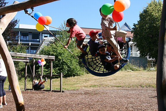 Gruppe von Kindern schaukelt gemeinsam auf einer großen Netzschaukel im Park. Einige halten bunte Ballons, lachen und bewegen sich schwungvoll. Sommerliches Wetter, fröhliche Spielszene.