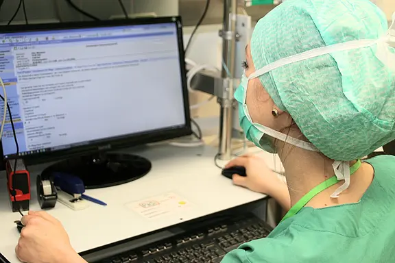 Medizinische Fachkraft in grüner OP-Kleidung und Maske sitzt am Klinikarbeitsplatz vor einem Monitor und bearbeitet eine elektronische Patientenakte mit Maus und Tastatur.
