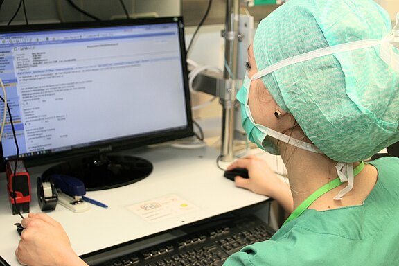 Medizinische Fachkraft in grüner OP-Kleidung und Maske sitzt am Klinikarbeitsplatz vor einem Monitor und bearbeitet eine elektronische Patientenakte mit Maus und Tastatur.