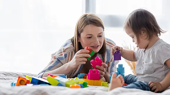 Eine erwachsene Person und ein Kleinkind liegen auf einem Bett und spielen mit bunten Steckbausteinen. Das Kind baut einen kleinen Turm, die Erwachsene unterstützt lächelnd.