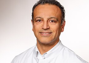 M. Shafie Ariai Ein lächelnder Mann trägt einen weißen Kittel. Sein Gesicht ist freundlich und er hat dunkle, leicht wellige Haare. Der Hintergrund ist schlicht und hell. Die Darstellung vermittelt einen professionellen Eindruck, der häufig mit medizinischen Berufen assoziiert wird.