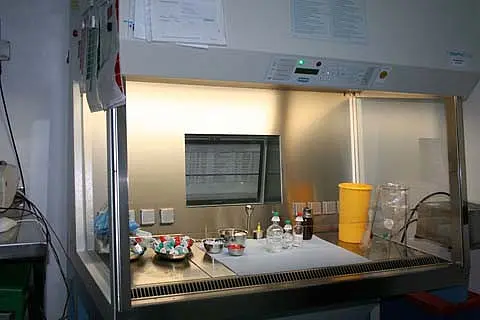 Ein Laborarbeitsplatz ist sichtbar, ausgestattet mit einem LED-beleuchteten Sicherheitswerkbank. Vor der Glasfront stehen mehrere Flaschen und Gläser auf einem Tisch. Im Hintergrund sind Notizen an der Wand und ein Bildschirm zu erkennen. Die Umgebung wirkt sauber und organisiert.