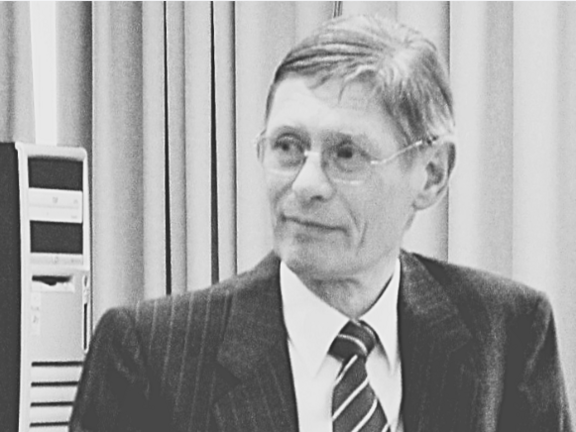 Schwarz-weiß-Foto: Ein älterer Mann im Anzug mit gestreifter Krawatte sitzt in einem Innenraum und blickt nach rechts. Links daneben steht ein Desktop-Computergehäuse. Neutrale, sachliche Szene.