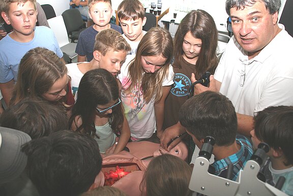 Gruppe von Schulkindern beobachtet gespannt eine medizinische Demonstration: Ein Ausbilder zeigt an einem Torso‑Simulator mit beleuchtetem Innenraum, wie chirurgische Instrumente eingesetzt werden. Die Kinder stehen eng um das Modell und lernen praktische Schritte.