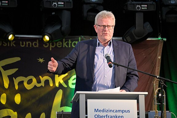 Älterer Mann mit Brille im Anzug spricht auf einer Bühne am Rednerpult. Vor ihm ein Mikrofon; er gestikuliert. Auf dem Pult steht „Medizincampus Oberfranken“. Bühnenbeleuchtung im Hintergrund.