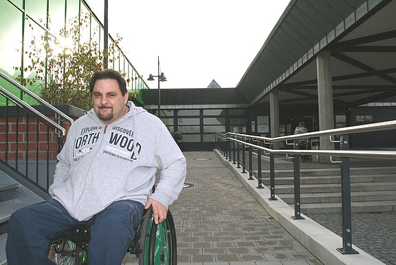 Ein Mann im Rollstuhl sitzt am Eingang eines modernen Gebäudes, neben einer breiten Rampe mit Geländern und Treppen. Die Szene betont barrierefreien Zugang.