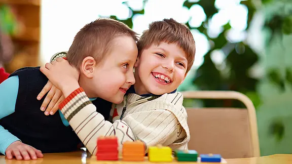 Zwei lachende Kinder sitzen an einem Tisch und umarmen sich herzlich. Vor ihnen liegen bunte Spielsteine. Die Szene vermittelt Freundschaft, Freude und Zusammenhalt.