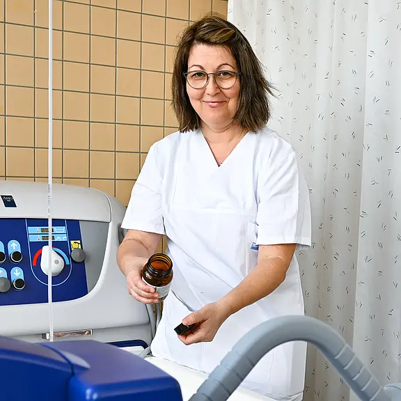 Eine Pflegekraft in weißer Uniform lächelt. Sie hält ein braunes Gläserchen in der einen Hand und einen Deckel in der anderen Hand. Im Hintergrund ist ein medizinisches Gerät zu sehen. Der Raum wirkt hell und ordentlich, mit einer Trennwand aus Vorhängen.