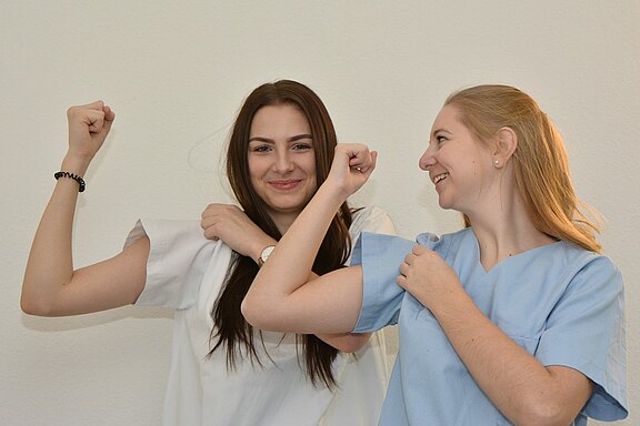 Zwei Frauen in medizinischer Kleidung zeigen lächelnd ihre starken Oberarme, während sie sich gegenseitig anlächeln. Die Atmosphäre ist freundlich und motivierend, in einem hellen Raum mit einer neutralen Wand im Hintergrund.
