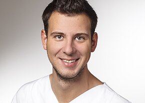 Station 21 Porträtfoto: junger Mann mit kurzen dunklen Haaren lächelt in die Kamera. Er trägt ein weißes V‑Oberteil vor neutralem, hellem Hintergrund. Fokus auf freundlichem Gesichtsausdruck.