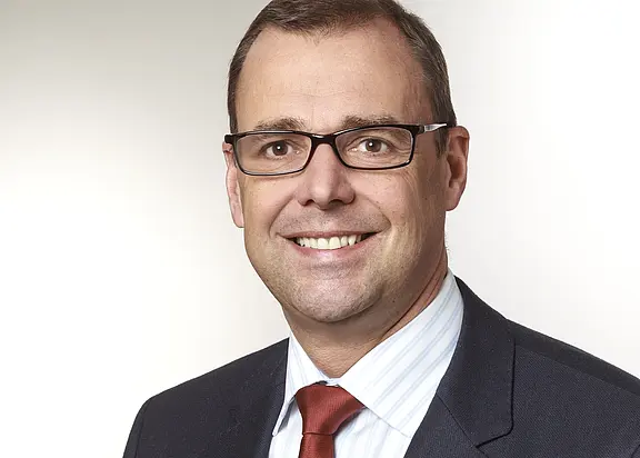 Professionelles Porträt: lächelnder Mann mit Brille, kurzen braunen Haaren, glatt rasiert. Er trägt dunklen Anzug, weißes Hemd und rote Krawatte vor hellem, neutralem Hintergrund.