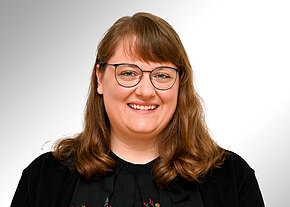 Lisa Lippert Porträtfoto einer lächelnden Frau mit Brille und schulterlangem braunen Haar vor neutralem, hellgrauem Hintergrund. Sie trägt eine schwarze Bluse und eine dunkle Strickjacke. Frontalaufnahme.