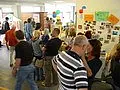 Eine Gruppe von Menschen steht in einem Raum, der mit bunten Plakaten dekoriert ist. Einige Personen sprechen miteinander, während andere Informationen an einer Wand lesen. Der Raum wirkt lebhaft und einladend.