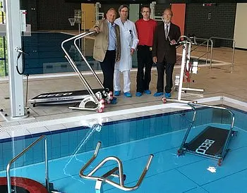Vier Männer stehen am Beckenrand eines Schwimmbades. Im Hintergrund sind verschiedene Geräte und eine Rampe zu sehen, die wahrscheinlich für die Unterstützung von Menschen mit eingeschränkter Mobilität gedacht sind.