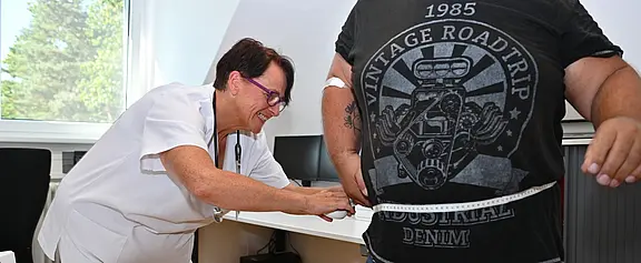 Pflegefachkraft in einer Praxis misst mit einem Maßband den Taillenumfang einer stehenden Person.