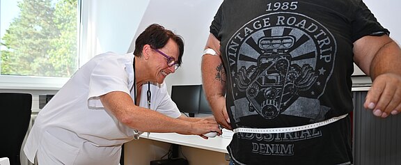Pflegefachkraft in einer Praxis misst mit einem Maßband den Taillenumfang einer stehenden Person.