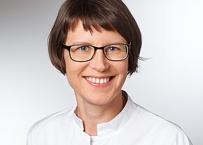 Porträt einer lächelnden Frau mit kurzem braunem Haar und schwarzer Brille, in weißem Kittel vor neutralem Hintergrund.