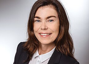 Professionelles Porträt: Eine lächelnde Frau mit schulterlangem braunem Haar trägt weiße Bluse und dunklen Blazer, vor neutralem Hintergrund, Blick zur Kamera.