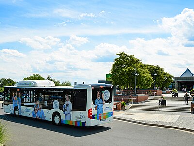 Ein Bus mit Werbung für eine Karriere im Gesundheitswesen fährt vorbei. Auf der Seite des Busses sind mehrere Personen in medizinischer Kleidung abgebildet. Der Himmel ist blau mit weißen Wolken, und im Hintergrund sind Gebäude und Bäume sichtbar.