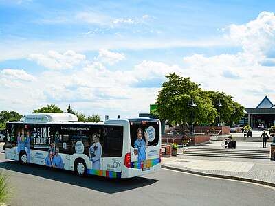 Ein Bus mit Werbung für eine Karriere im Gesundheitswesen fährt vorbei. Auf der Seite des Busses sind mehrere Personen in medizinischer Kleidung abgebildet. Der Himmel ist blau mit weißen Wolken, und im Hintergrund sind Gebäude und Bäume sichtbar.