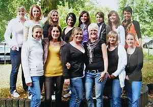 Eine Gruppe von 15 Frauen lächelt vor einem grünen Hintergrund. Sie stehen in mehreren Reihen und tragen verschiedene Kleidungsstile. Die Stimmung wirkt freundlich und fröhlich, und die Frauen scheinen sich gut zu verstehen.