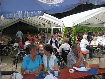 Eine Gruppe von Menschen sitzt unter Zelten in einem Garten. Einige Personen nutzen Rollstühle. Es gibt Tische mit Getränken, und die Atmosphäre wirkt gesellig und einladend. Menschen unterhalten sich und genießen die Zeit miteinander.
