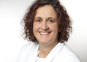 Susanne Dietze Porträt einer lächelnden Frau mit lockigem, kastanienbraunem Haar, die einen weißen Arztkittel trägt. Der Hintergrund ist hell und neutral. Die Frau strahlt Freundlichkeit und Professionalität aus.