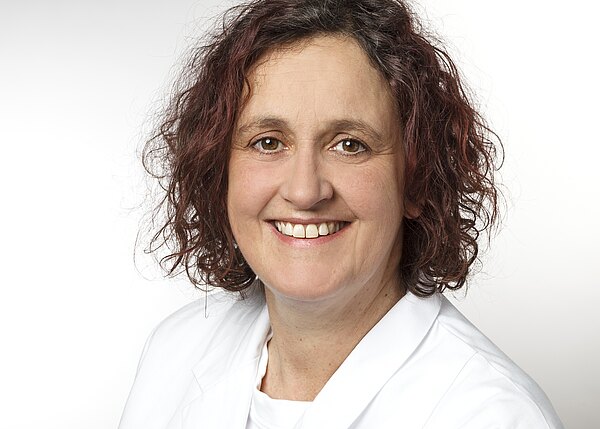 Porträt einer lächelnden Frau mit lockigem, kastanienbraunem Haar, die einen weißen Arztkittel trägt. Der Hintergrund ist hell und neutral. Die Frau strahlt Freundlichkeit und Professionalität aus.