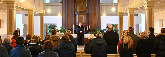 Innenraum einer Kirche: Eine Gemeinde steht und blickt nach vorn, wo ein Geistlicher in schwarzer Robe am Altar mit Kerzen predigt.