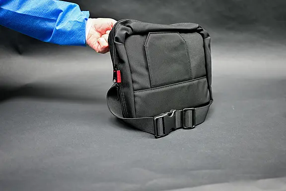 Tasche für Kunstherz-Equipment Hand in blauer Jacke hält eine kompakte, schwarze Tasche mit breitem, verstellbarem Gurt und Steckschließe. Die Tasche wirkt gepolstert und robust.