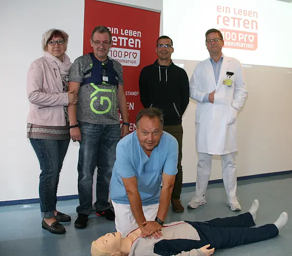 Übung zur Wiederbelebung: Ein Mann kniet am Boden und führt Herzdruckmassagen an einer Reanimationspuppe durch. Hinter ihm beobachten mehrere Personen, darunter ein Arzt im weißen Kittel. Plakat im Hintergrund: „Ein Leben retten – Reanimation“.