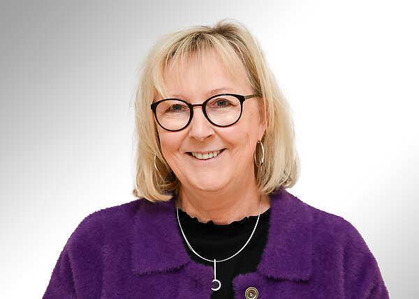 Porträt einer lächelnden Frau mit Brille und schulterlangem blondem Haar. Sie trägt eine lila Jacke, ein schwarzes Oberteil und eine silberne Halskette. Heller, neutraler Hintergrund.