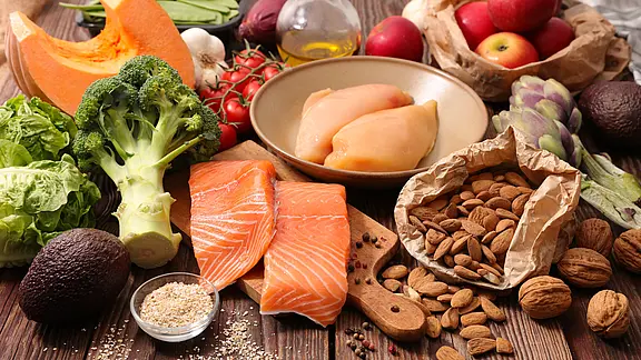 Gesunde Lebensmittel auf Holzuntergrund: zwei Lachsfilets und Hähnchenbrust, umgeben von Brokkoli, Avocado, Kürbis, Salat, Kirschtomaten, Artischocke, Äpfeln, Mandeln, Walnüssen, etwas Getreide und Olivenöl. Darstellung einer ausgewogenen, frischen Ernährung.