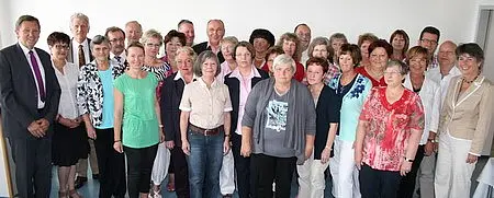 Eine Gruppe von etwa 40 Menschen steht in einem Raum. Die Mehrheit sind ältere Frauen, die in verschiedenen Farben gekleidet sind. Einige Männer sind ebenfalls anwesend. Alle schauen in Richtung der Kamera und lächeln. Der Hintergrund ist hell und neutral.