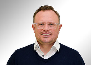 Porträt eines lächelnden Mannes mit kurz geschnittenen Haaren, heller Kunststoffbrille, weißem Hemd und dunkelblauem Pullover, frontal vor neutralem, hellem Hintergrund.