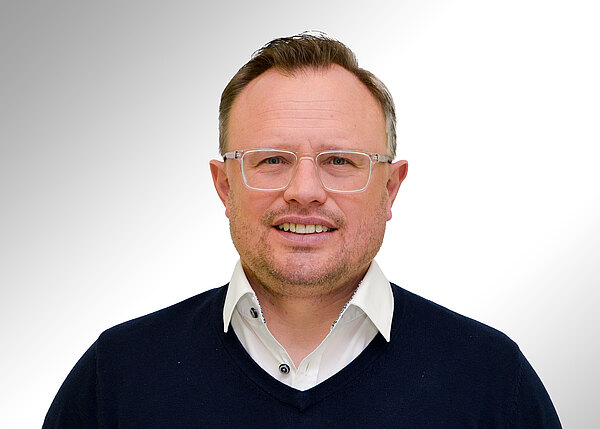 Porträt eines lächelnden Mannes mit kurz geschnittenen Haaren, heller Kunststoffbrille, weißem Hemd und dunkelblauem Pullover, frontal vor neutralem, hellem Hintergrund.