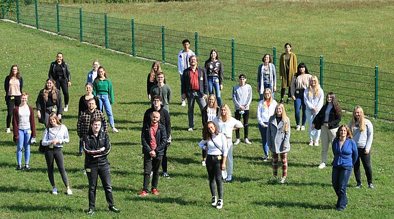Eine Gruppe von ungefähr 30 Personen steht auf einer Wiese, umgeben von einem Zaun. Die Menschen lächeln und posieren in freundlicher Atmosphäre. Es sind sowohl Männer als auch Frauen in unterschiedlichen Kleidungsstilen und Altersgruppen vertreten. Die Sonne scheint und der Hintergrund ist grün.
