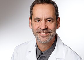 Dr. med. Sebastian Ortlieb Porträt eines lächelnden Mannes mittleren Alters in weißem Laborkittel vor neutralem Hintergrund; freundlicher, professioneller Ausdruck.