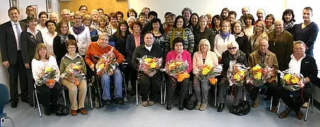 Gruppenfoto mit etwa 50 Personen, die überwiegend sitzen. Viele der Anwesenden halten bunte Blumensträuße in den Händen. Einige stehen im Hintergrund. Die Atmosphäre wirkt festlich und freundlich, wahrscheinlich anlässlich eines besonderen Ereignisses oder einer Feier.