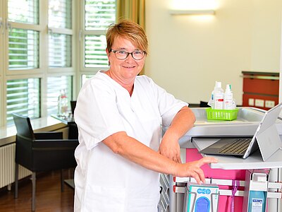 Pflegefachkraft in weißer Arbeitskleidung steht an einem Pflegewagen mit Laptop und Desinfektionsmitteln in einem hellen Klinikraum und lächelt in die Kamera.