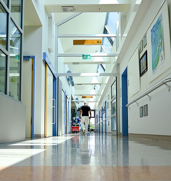 Heller, langer Krankenhausflur mit glänzendem Boden; Tageslicht fällt durch Oberlichter. Eine einzelne Person geht in der Ferne den Gang entlang, Richtung Ausgang. Türen und Wandschilder säumen den Flur.